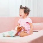 Criança sorrindo sentada na Cama Montessoriana Aconchego rosa da Keiko, 100% espuma e bordas macias, em quarto infantil claro e seguro.