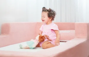 Criança sorrindo sentada na Cama Montessoriana Aconchego rosa da Keiko, 100% espuma e bordas macias, em quarto infantil claro e seguro.